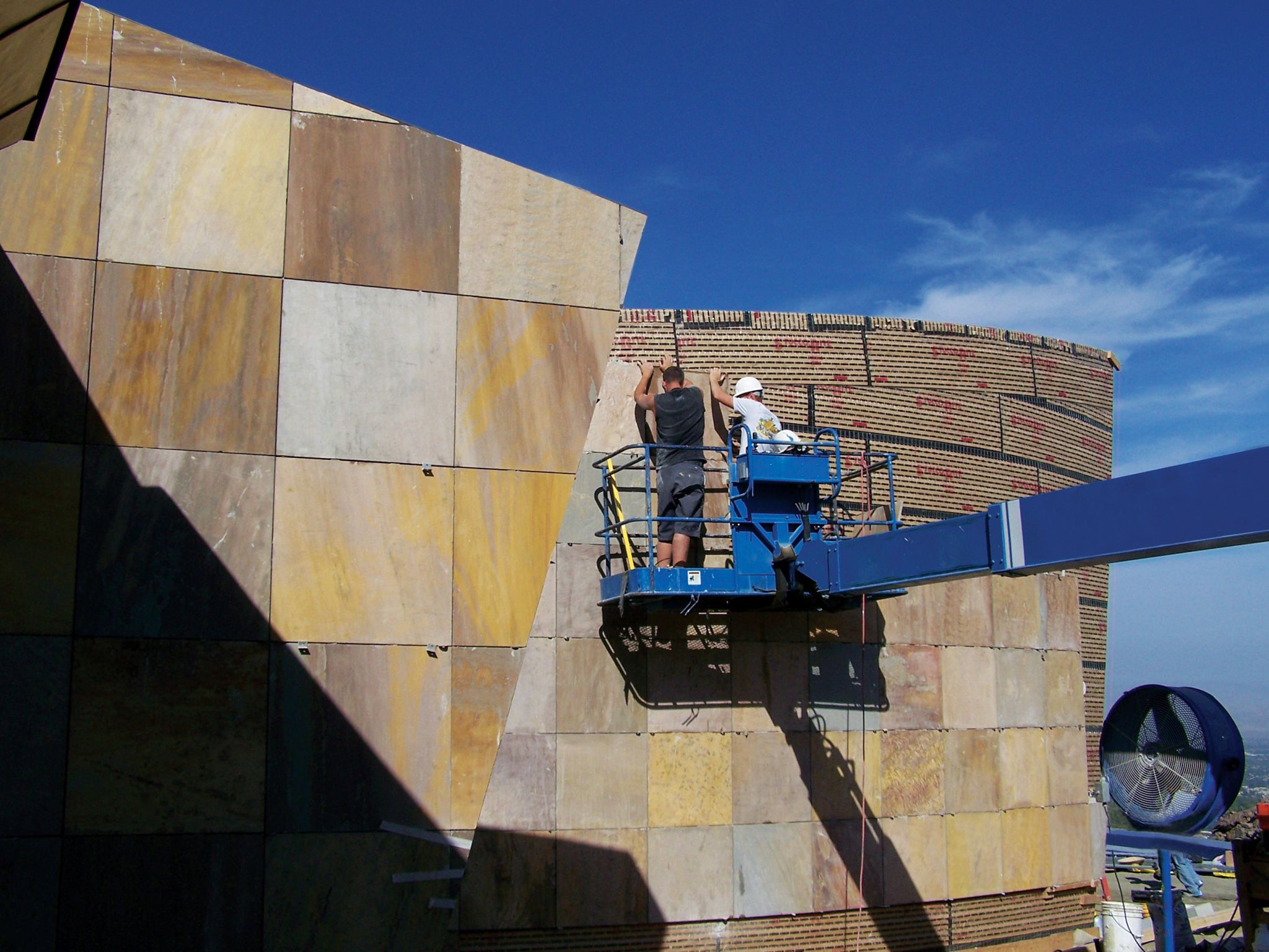 These workers are using a manlift to mechanically anchor sandstone to a building’s exterior wall with metal angle clips.