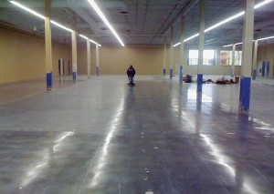 The special silica-polymer dispersion stain protector was applied, and the floor burnished again. To the left of the operator, the stain-protected, unburnished surface is seen as a dull, matte grey. To his right and in the foreground, the burnished surface shows strong reflectivity, visually the equivalent gloss of an 800-grit polish on a surface that would otherwise not be polishable.