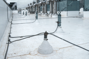 UL-listed through-structure penetration, in foreground, is used to connect rooftop conductors to down-conductors inside the building. The flashing used to seal the penetration can be a type typically specified with the roofing. Cables lead to parapet-mounted air terminals (on left) and fume hood exhaust fans (on right). Window washing equipment anchors are bonded to the lightning protection system and also brace the tall exhaust fans. Photo courtesy Boston Lightning Rod Company 