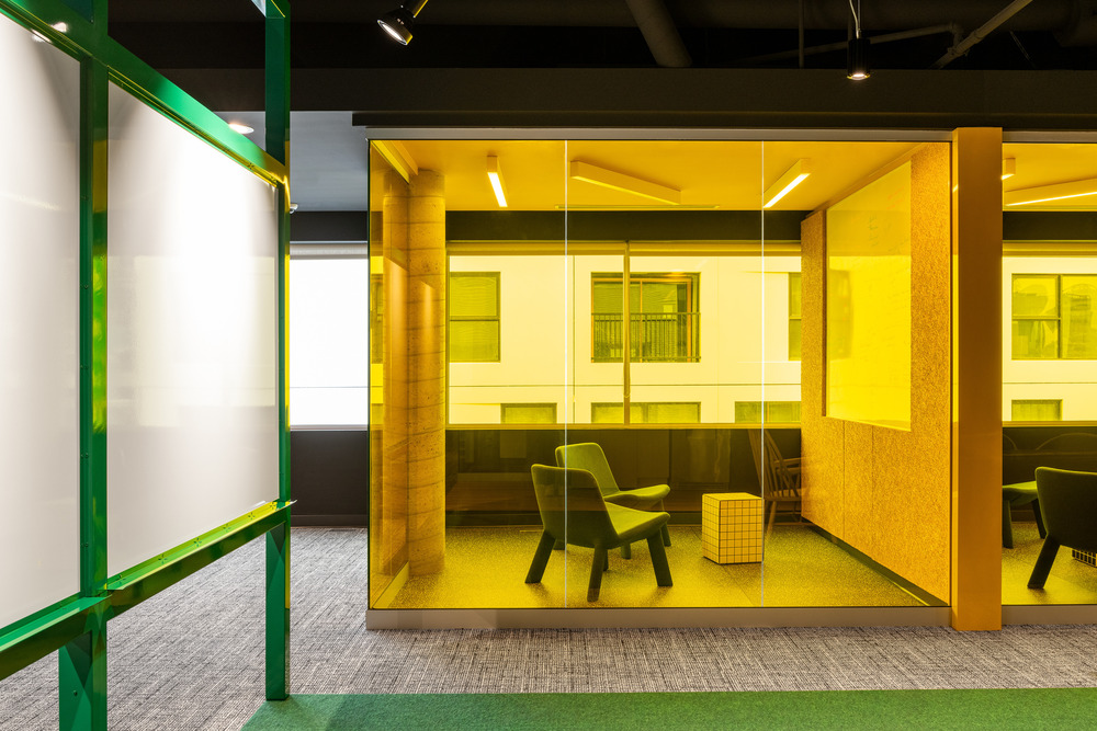 A private employee room is enclosed by a neon-yellow glass, while in the foreground a bright-green colored common area is visible.