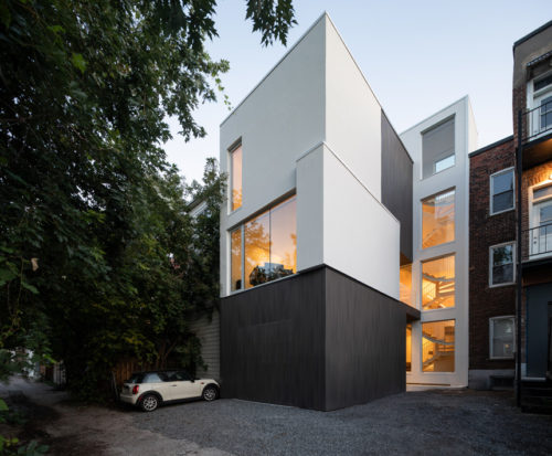 Modern triplex in Montreal with contrasting dark and light facades, large windows, and a visible interior staircase at dusk.