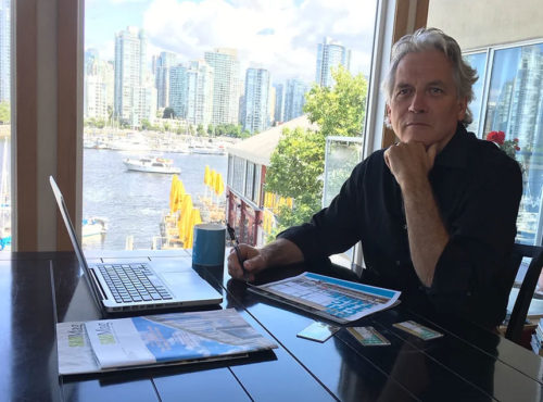 a man with long white hair sitting with a laptop with a waterfront in the background