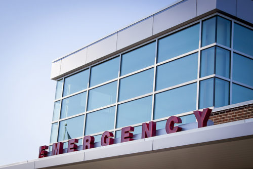 Emergency room entrance with red block letters on the metal awning of a hospital building, blue tone filter.