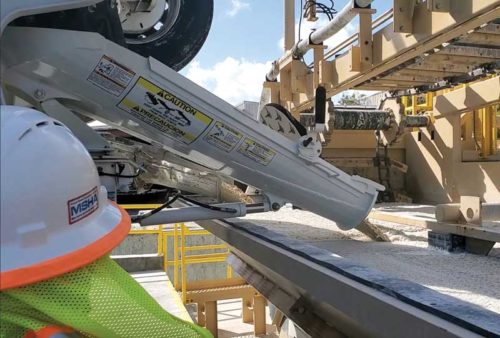 A construction worker in safety gear monitors concrete being poured onto a conveyor system at a batching or precast facility.