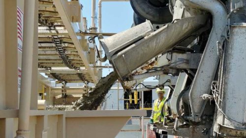 A concrete mixer truck pours fresh concrete into a batching or precast system, with a worker in safety gear overseeing the process.