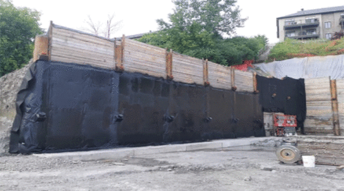 Construction site with a retaining wall reinforced by black waterproofing material, wooden planks, and steel braces, surrounded by gravel and construction equipment, with trees and residential buildings in the background.