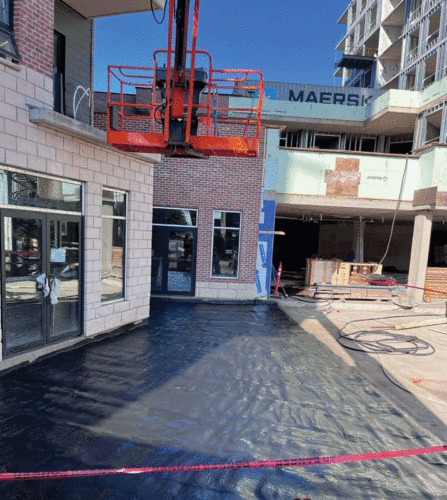 Construction site featuring a partially completed building with brick and stone facades, a black waterproof membrane covering the ground, and a red aerial lift platform in the foreground. Surrounding structures and construction materials are visible under a clear blue sky.