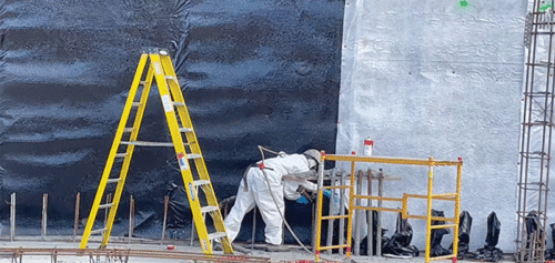 Construction worker in protective white coveralls applying material to a wall covered with black and white waterproof membranes, with a yellow ladder and safety scaffolding in the foreground.