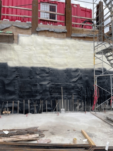 Construction site featuring a retaining wall coated with black and white waterproof membranes, reinforced with wooden supports and scaffolding. The site includes exposed rebar, wooden planks, and a red shipping container on the upper level.