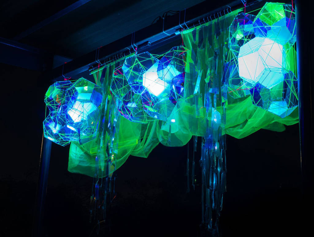 An illuminated geometric art installation at DesignTO Festival, glowing in blue and green hues with draped translucent fabric.