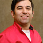 A smiling Ali Ahrabi is posed in front of a neutral-toned background. He wears a bright red shirt, unbuttoned at the collar, over a white undershirt. His dark hair is styled neatly, and he appears confident and approachable.