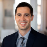 Alex Ardelean smiles warmly at the camera, wearing a dark suit with a patterned tie. He has short, neatly styled hair and is positioned in an office environment with blurred background elements suggesting a bright, professional setting.