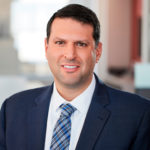 Peter Babaian with short dark hair is wearing a navy suit and a light blue checkered tie. The background shows a softly blurred office environment, suggesting a professional setting.