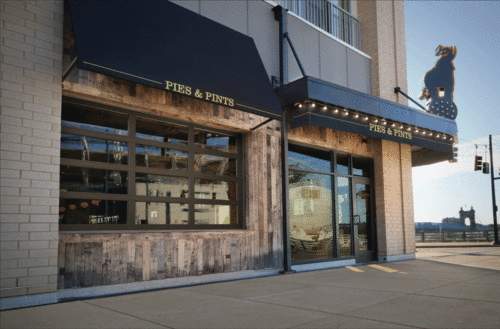 The exterior of a Pies & Pints restaurant, featuring a modern storefront with vertically stacking glass doors, a rustic wooden facade, and black awnings with the restaurant name displayed.