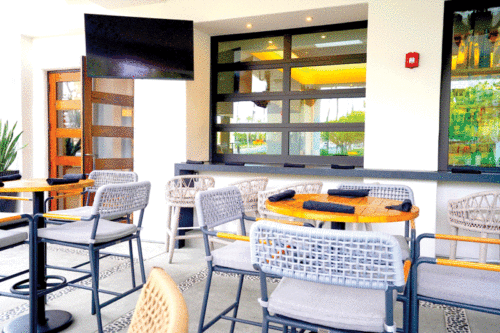 Outdoor patio with woven chairs, round wooden tables, a wall-mounted TV, and a large window. Bright, inviting atmosphere with modern decor.