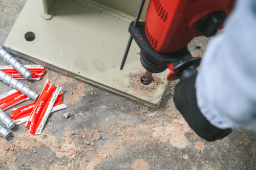 A person holding a red drill is creating a hole in a green metal surface. Nearby, several silver attachments and small red packaging are scattered on the ground, along with wood shavings from the drilling process.