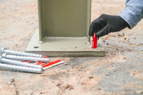 A hand wearing a black glove is positioning a red object against a green metal base anchored on a sandy surface. Several silver screws are scattered nearby.