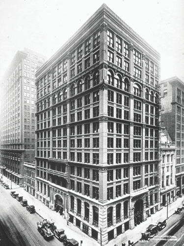 A historic photograph of a large, multi-story building situated at a city intersection. The building features a decorative façade with arched windows and intricate architectural details. Several vintage automobiles are parked along the street in front of the building, indicating an early 20th-century setting.