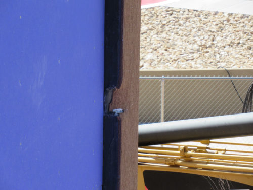 The image depicts the corner of a wall painted purple. The paint is peeling, revealing the brown wood underneath. In the background, there is a construction area with a yellow piece of machinery, and a fence is visible.