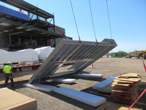 A construction worker in a bright yellow shirt stands on a gravel surface, guiding a large, angled structure being lifted by cranes at a construction site. The structure has a metallic frame and is partially obscured by white material. In the background, an incomplete building and stacked materials are visible under a clear blue sky.