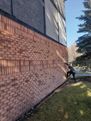 A person is climbing a sloped brick wall beside a building. The wall is made of red bricks, and the background features grass and trees, indicating an outdoor setting. The individual is dressed in a gray coat and appears focused on reaching higher on the wall.