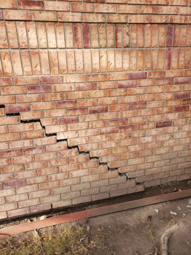 A brick wall displays significant cracking, with a diagonal section shifting downwards, revealing a gap at the base. The bricks have various shades of red and brown, and a metal beam lays on the ground below.