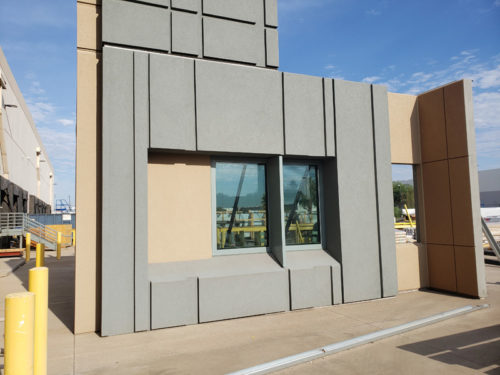 The image shows a modern architectural structure featuring a large window at the center of a corner. The exterior walls are a combination of gray and beige panels, creating a geometric pattern. The setting includes a clear blue sky in the background.