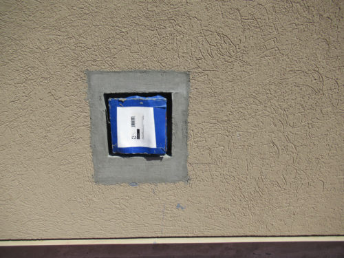 A textured beige wall features a square cutout surrounded by gray tape. Inside the cutout, a blue object is partially visible, held in place with blue tape, displaying what appears to be a white label with black text.