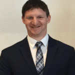 Mark D. Hagel, smiling, wearing a navy-blue suit and a light blue and gray plaid tie. He has short, dark hair and is standing in front of a plain light-colored background.