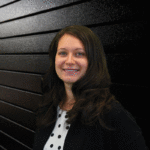 Professional headshot of Heather Bender with long dark hair, wearing a black blazer and a white polka-dot blouse, smiling in front of a textured black background.