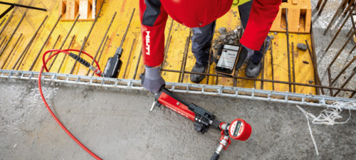 A construction worker wearing a red and black jacket operates a Hilti tool to secure rebar on a concrete surface. A tablet rests on the ground, showing measurements or data related to the task. The background features construction materials and equipment.