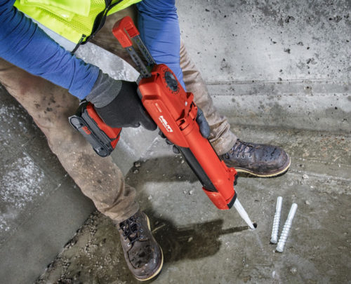 A construction worker in a bright yellow safety vest and blue shirt is using a red caulking gun. They are positioned in a concrete area, with three screws lying on the ground nearby. The worker is wearing gloves and sturdy boots, indicating they are engaged in a construction or repair task.