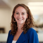 Elizabeth Rodenkirch with curly brown hair is wearing a blue blazer and large hoop earrings. She is positioned against a softly blurred office background, suggesting a professional environment.