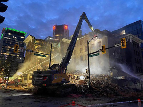 Nighttime demolition scene of a multi-story building, with an excavator operating amidst rubble and debris. Bright spotlights illuminate the structure and water hoses spray the site, likely for dust control. Surrounding buildings with lit windows and a dark, cloudy sky are visible in the background.