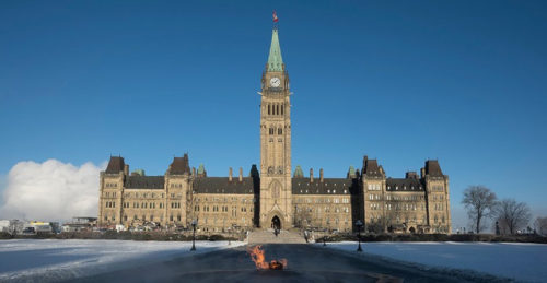 A view of Parliament Hill in Ottawa during winter. The iconic clock tower stands tall against a clear blue sky, while snow blankets the ground. In the foreground, there’s a small flame on the pathway, and the building's intricate architectural details are visible.