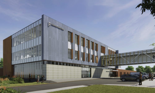 Modern three-story building with a sleek design featuring large windows, metal paneling, and wooden accents. A glass-enclosed skybridge connects to another structure. The building is surrounded by greenery, with people walking and cars parked in the foreground.