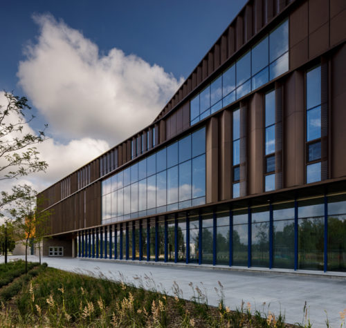 The image shows a modern building with a sleek, brown exterior and extensive glass windows reflecting the sky. In the foreground, there is a landscaped area with plantings alongside a paved pathway, contributing to the contemporary aesthetic of the structure.