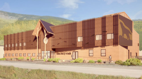 A contemporary wooden building stands in the foreground, set against a backdrop of mountains. The structure features an artistic representation of a wolf on its side, gabled roof with a central circular element, and several large windows. A pathway leads up to the entrance, where a few people are seen walking.