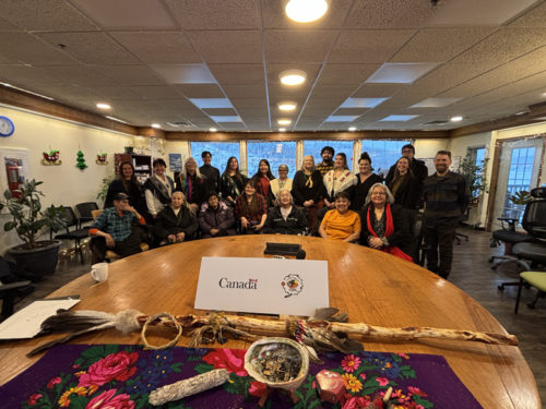 A diverse group of individuals is gathered around a circular wooden table in a brightly lit room. The atmosphere is warm and welcoming, with plants and decorations visible in the background. A sign at the center of the table reads "Canada," accompanied by Indigenous symbols. Various items are displayed on the table, including a decorated cloth with floral patterns, a ceremonial smudge bowl, and a walking stick. The people are smiling and are dressed in a mix of traditional and contemporary attire.