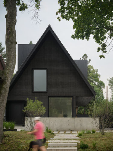 Modern house with a steep dark gabled roof, large windows, and a minimalist design surrounded by greenery, featuring a concrete base and a pathway leading to the entrance.