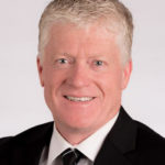 Professional headshot of Michael Hensen with short, light-colored hair, wearing a black suit and tie, smiling at the camera.