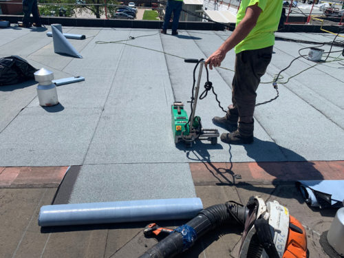 Worker in a neon shirt using a welding machine to install a gray roofing membrane on a flat roof, with tools and materials scattered around.