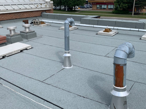 Rooftop view showing metal pipes, ventilation systems, and flat roofing material.