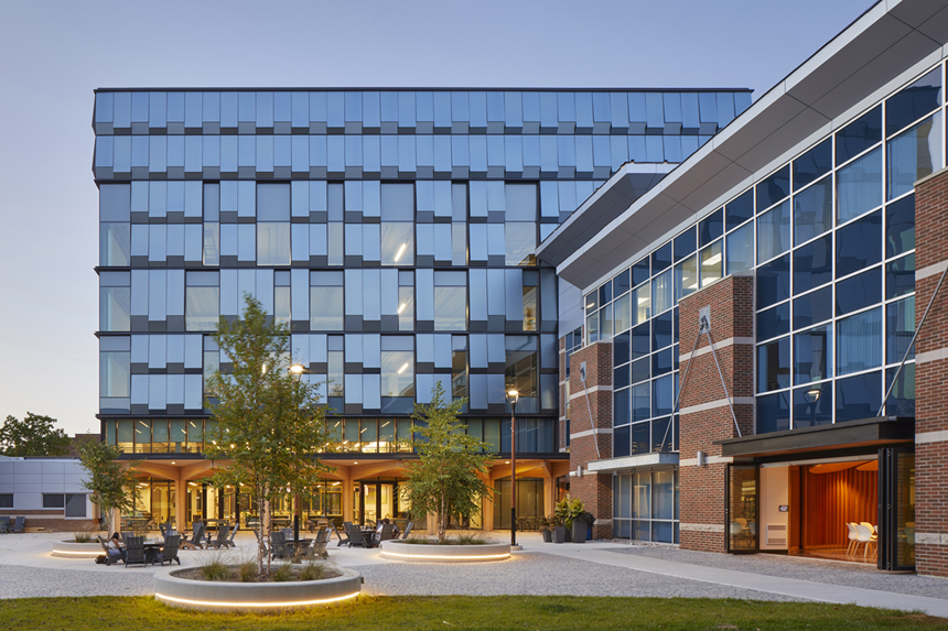 Modern building complex with glass and brick facades, featuring an outdoor courtyard with seating, trees, and illuminated planters at dusk