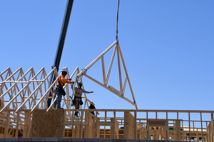 Construction workers are installing a wooden truss on a building site, with a crane lifting the truss into position against a clear blue sky. 