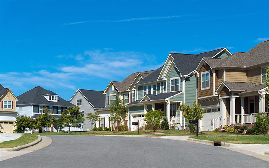 A suburban neighborhood with modern, colorful two-story houses, well-maintained lawns, and a clear blue sky.