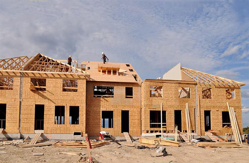 Partially constructed residential building with exposed wooden framing and workers installing the roof under a clear sky