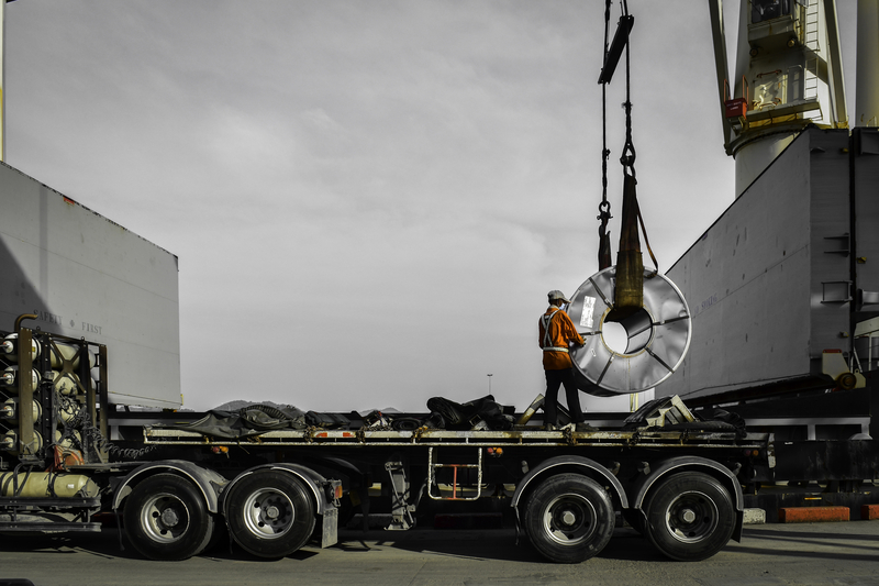 Steel coil being lifted off a transport truck. A construction worker is on the truck assisting with the load.