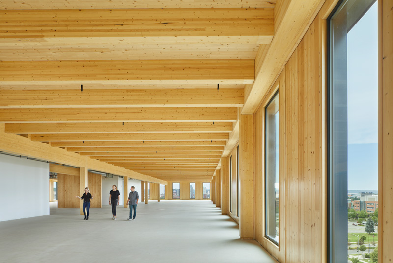 Three individuals walk through a spacious, modern interior featuring wooden beams and large windows, highlighting natural light and simplicity.