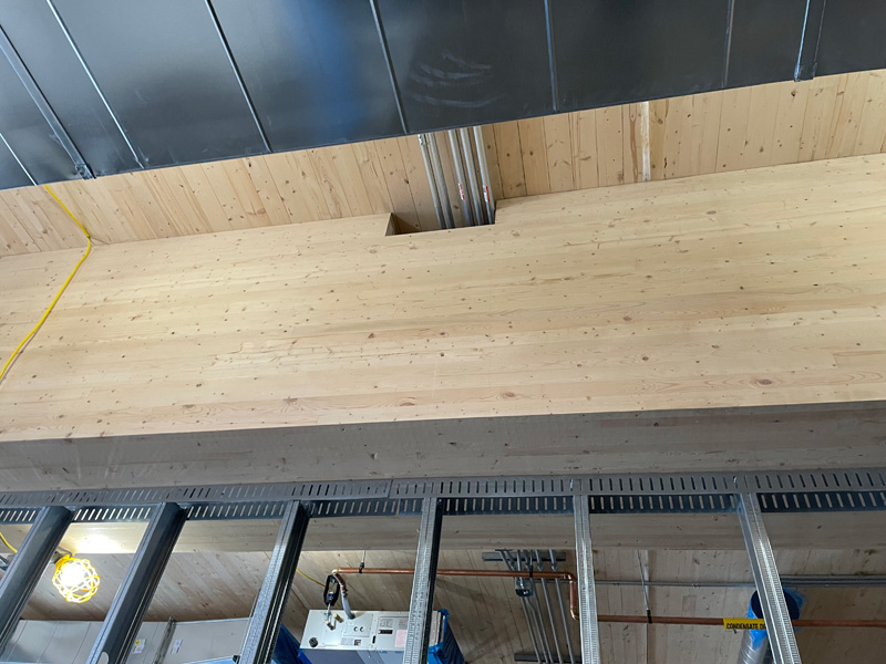 View of a wooden ceiling with metal beams and exposed pipes, highlighting construction details and overhead electrical wiring.